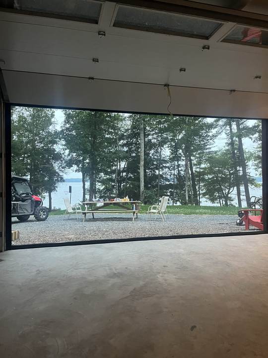 View from inside a garage toward a lake, trees, a picnic table and an off road vehicle